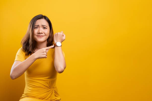 A woman in a yellow dress keenly pointing at her watch in front of a vibrant yellow background.