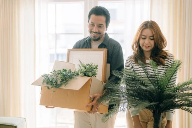 Newly-welcomed Asian couple enhancing tenant satisfaction by moving into a new home while carrying boxes and plants.