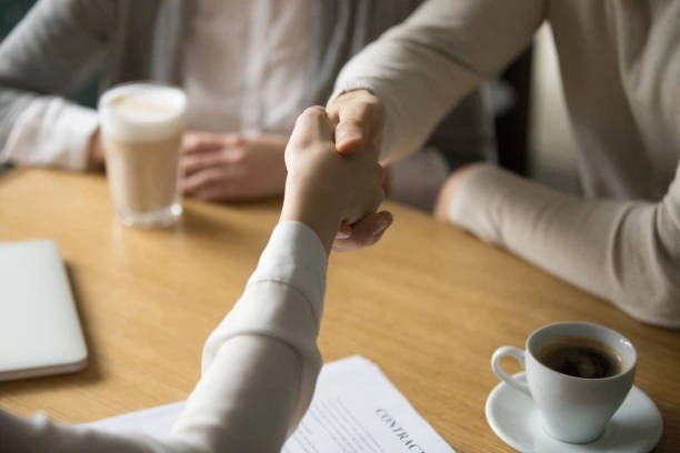 Two people shaking hands at a table to negotiate an outsource agreement for property management.