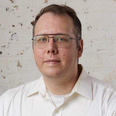 Portrait of a middle-aged man with glasses, representing the best property management company, wearing a white shirt against a textured, off-white background.