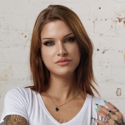 A woman with tattoos, wearing a white t-shirt and a necklace, poses against a brick wall background for the best property management company's advertising campaign.