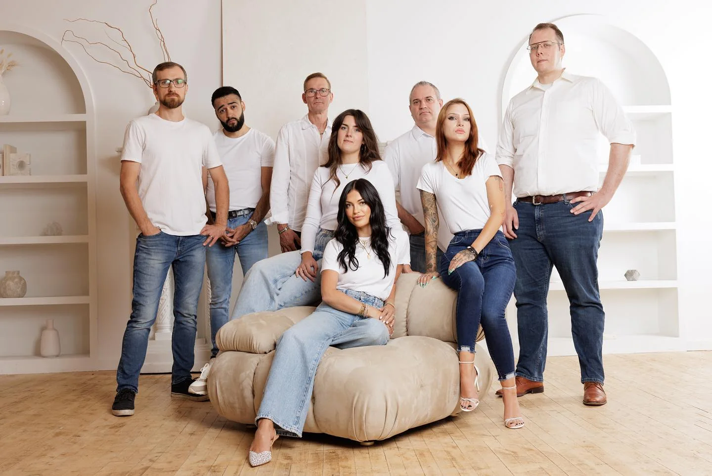 Group of eight diverse adults in white shirts and jeans, posing in the bright office of the best property management company, with one woman seated on a beige bean bag.