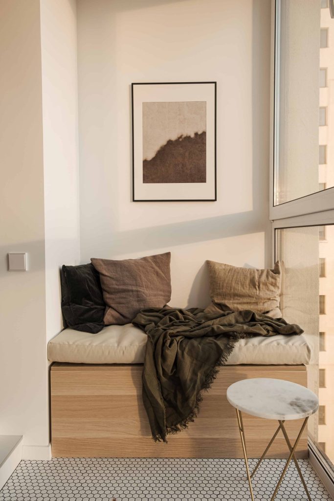 A cozy modern nook with a cushioned bench, throw pillows, a blanket made from sustainable materials, and a small round side table, next to a window with sunlight filtering through.