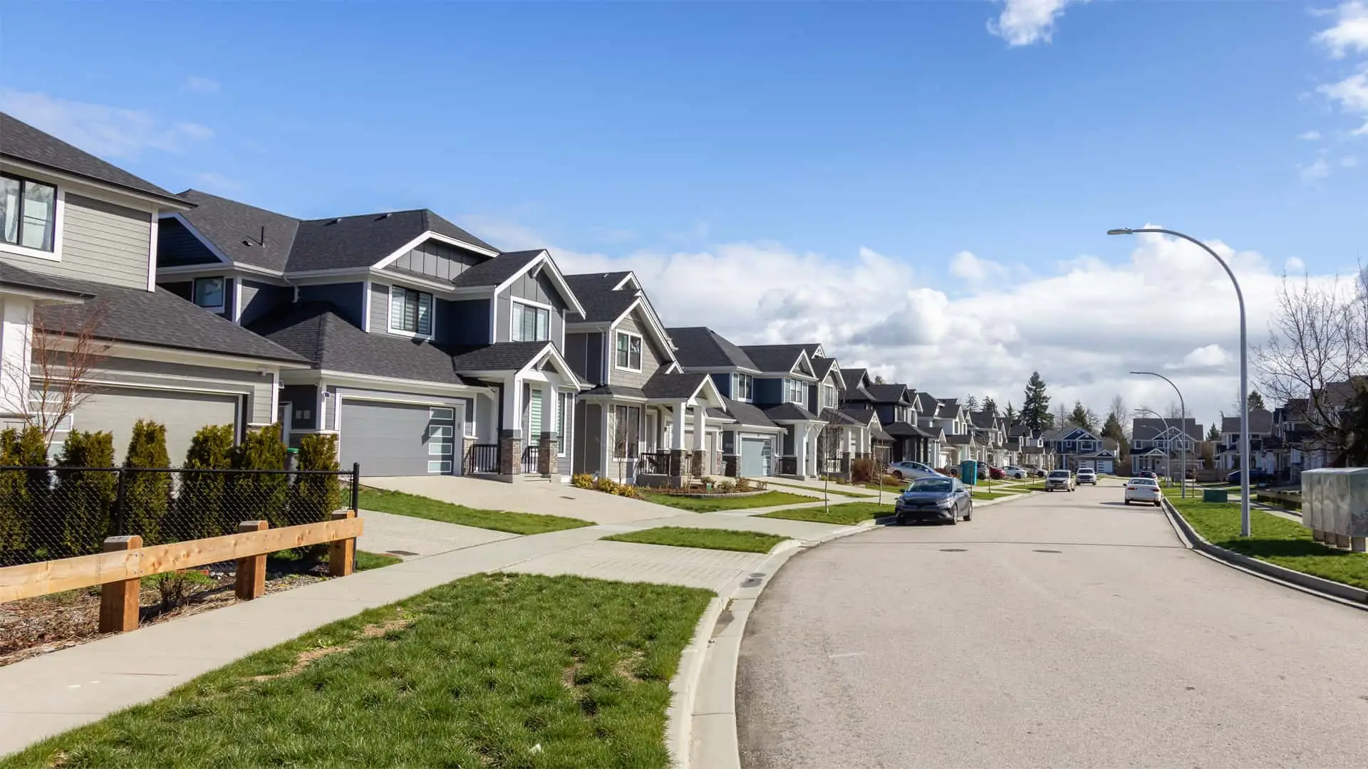 A suburban neighborhood with rows of modern two-story houses, neatly trimmed lawns, parked cars, and a clear blue sky, resembling the orderly charm you'd expect from the best property management in London.