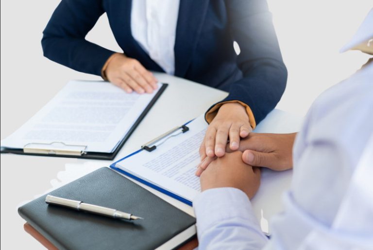 Two professionals sitting at a desk, one comforting the other by holding hands, with documents and a clipboard in front of them.