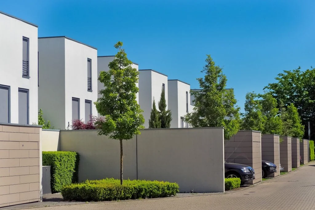 Modern row of white, cube-shaped townhouses boasts exceptional curb appeal with trees and parked cars lining a clean, quiet street under a clear blue sky.