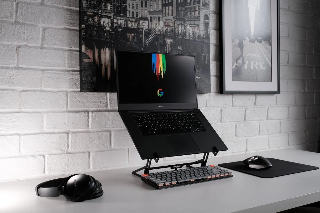 Laptop on a stand with a colorful Google logo on the screen, accompanied by a keyboard, mouse, mouse pad, and headphones on a white desk against a white brick wall with black and white framed art. Ideal setup for rentals for remote work.