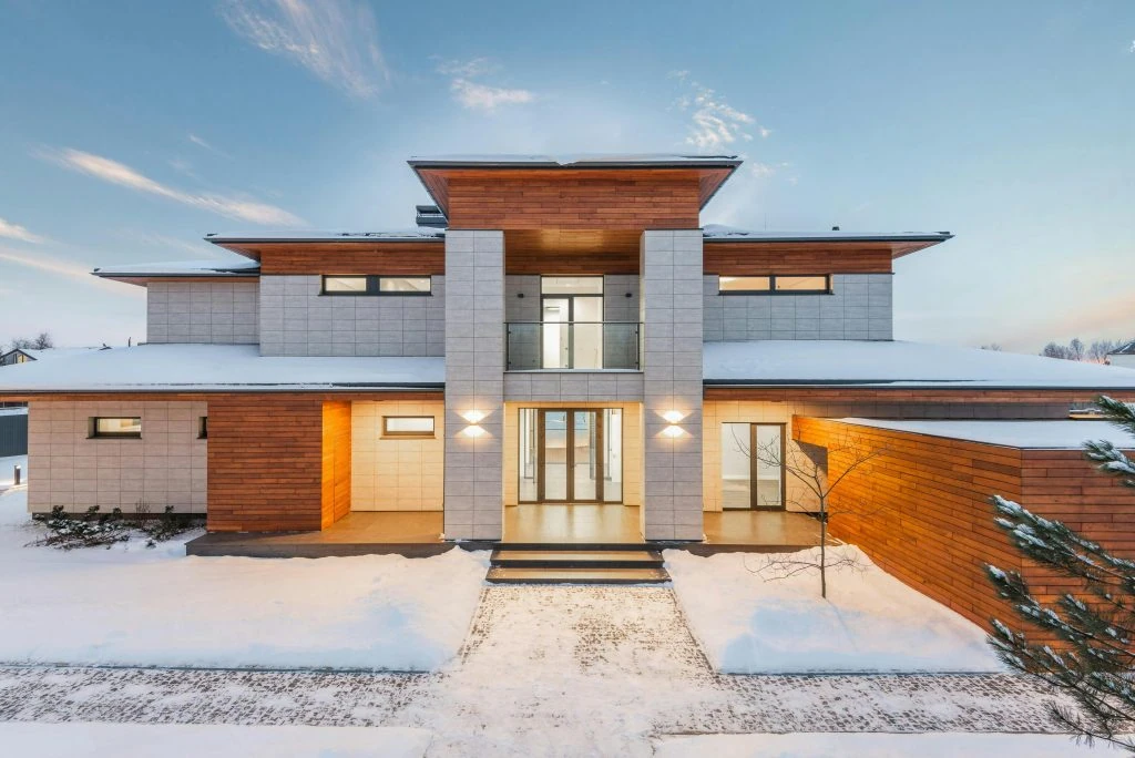 A modern, two-story house with a blend of wood and concrete exterior, featuring large windows, warm lighting, and surrounding snow-covered grounds reminiscent of the design principles promoted by Bill 163.