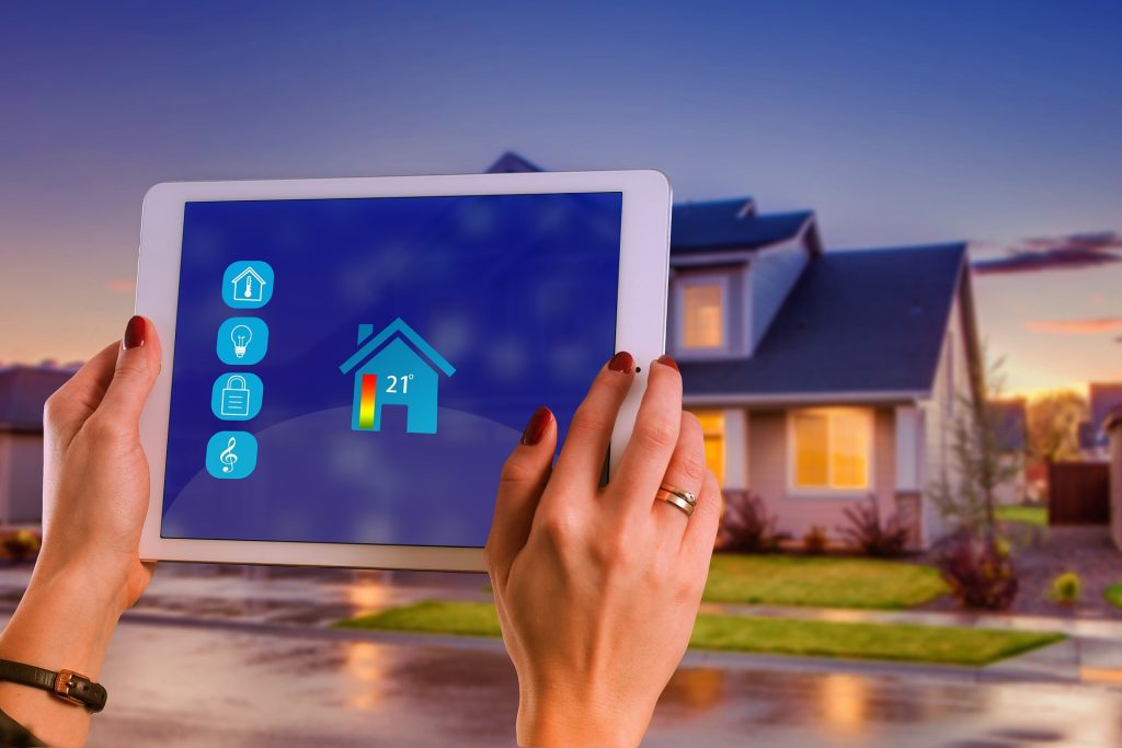 Hands holding a tablet displaying a home automation interface, with a modern house in the background at sunset, showcasing the future of property management.