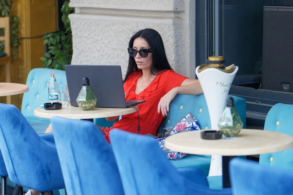 A person in a red dress and sunglasses sits at an outdoor café table, working on a laptop and exploring rentals for remote work.