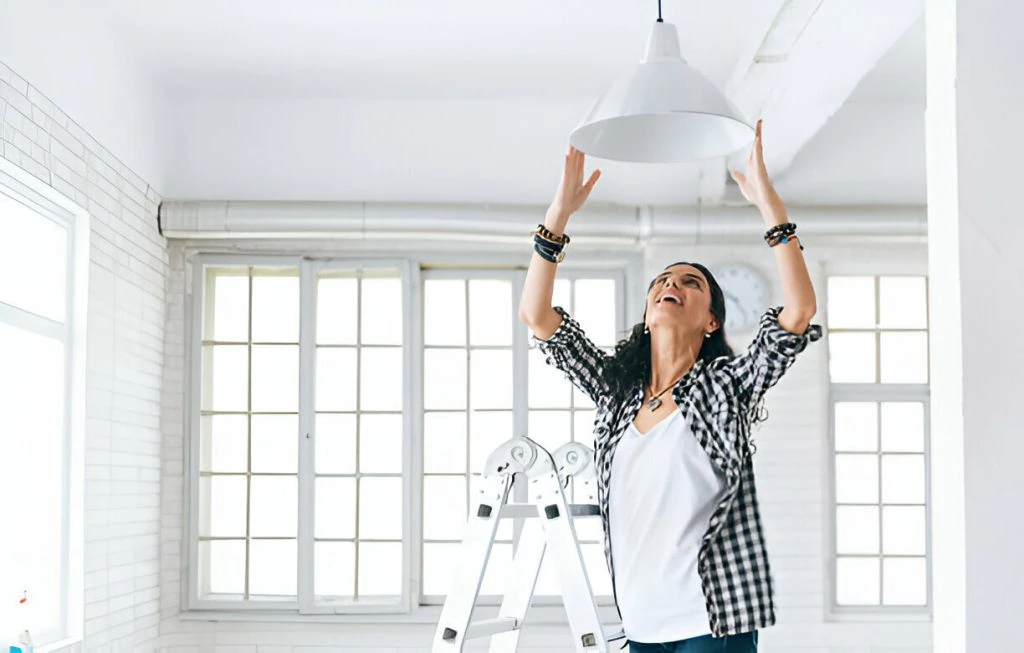 A person in a checkered shirt stands on a ladder, reaching up to adjust a hanging light fixture in a bright room with large windows, an essential step in attracting high-quality tenants, advanced property management, Discover advanced property management and maintenance strategies to attract high-quality tenants with strategic enhancements and effective marketing efforts for your rental property