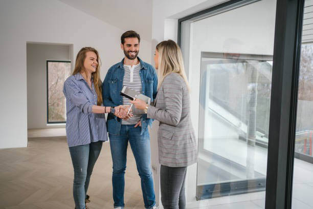 A couple stands in an empty room with large windows, shaking hands with a woman holding a tablet, perhaps discussing the recent implications of Bill 163.