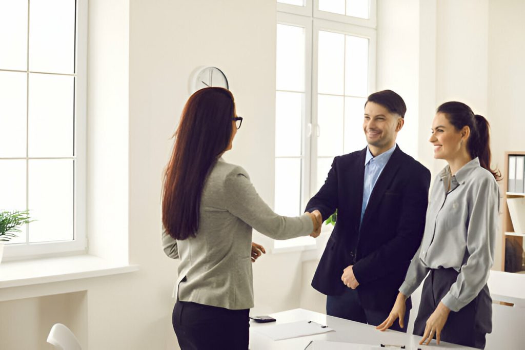 Three people in a bright office space. Two women, one with dark hair and glasses, and one with light hair, smile while shaking hands. A man in a dark blazer stands beside them, highlighting the positive landlord-tenant relationship being formed.