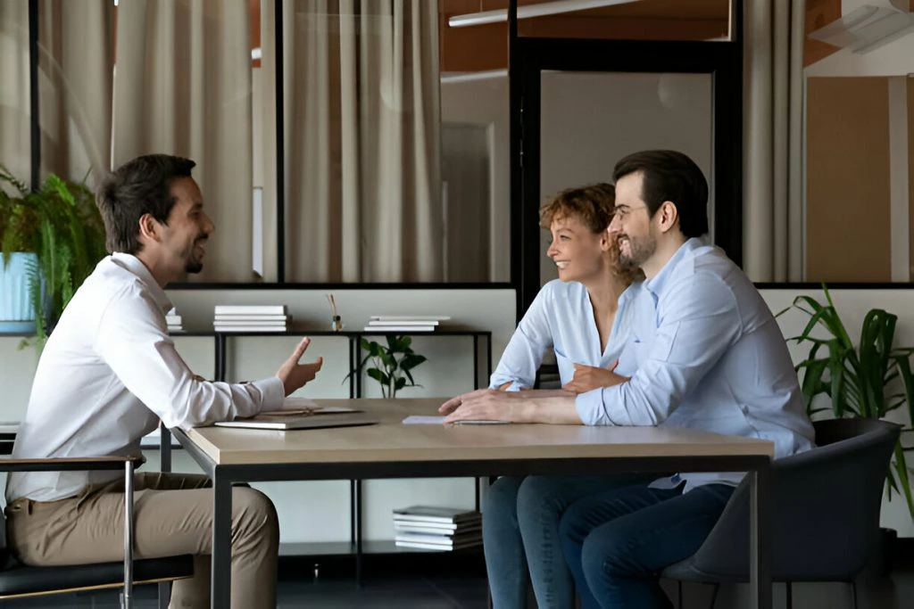 Three people engaged in a discussion around a table in an office setting, with two seated on one side and one on the other, strategizing ways to attract high-quality tenants against a backdrop of plants and shelves, Discover advanced property management and maintenance strategies to attract high-quality tenants with strategic enhancements and effective marketing efforts for your rental property