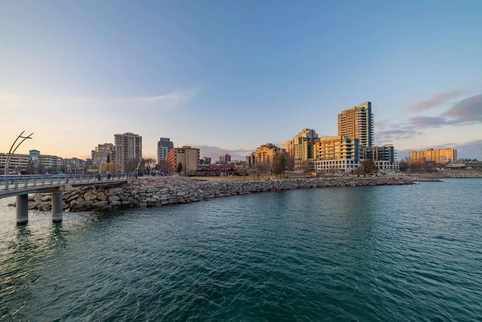 A waterfront view of a cityscape with modern high-rise buildings and a pier, captured at sunset with a clear sky and calm water—a perfect backdrop for property management Burlington.