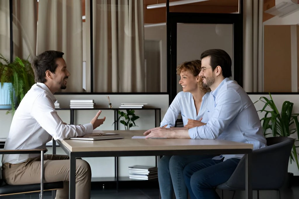 Three people are seated at a table in an office setting. A man on the left is talking, while a man and woman on the right are listening and smiling. Shelves and plants are visible in the background.