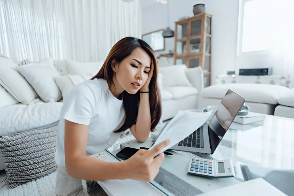 A woman sits on the floor by a table, looking at documents in her hand. She has a laptop, smartphone, and calculator in front of her. The room is bright and furnished with white furniture, creating a serene atmosphere as she analyzes rental market fluctuations, Property Management Service