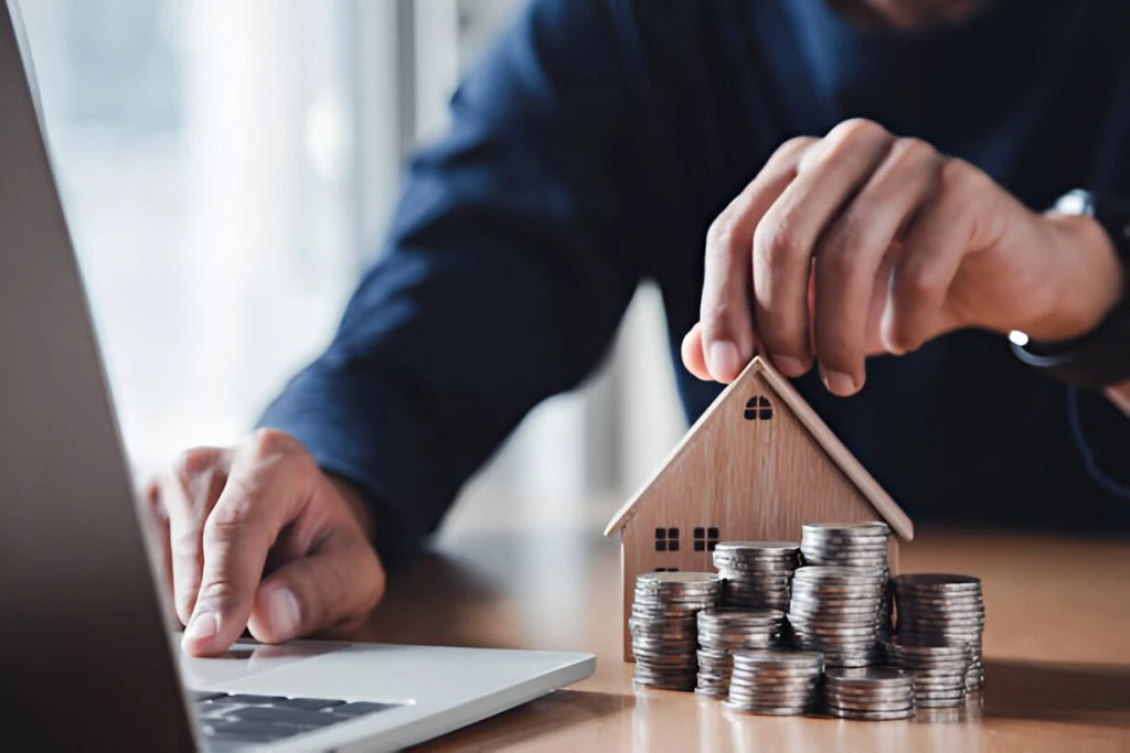 A person working on a laptop places a small wooden house model atop stacks of coins on a table, perhaps analyzing rental market fluctuations.