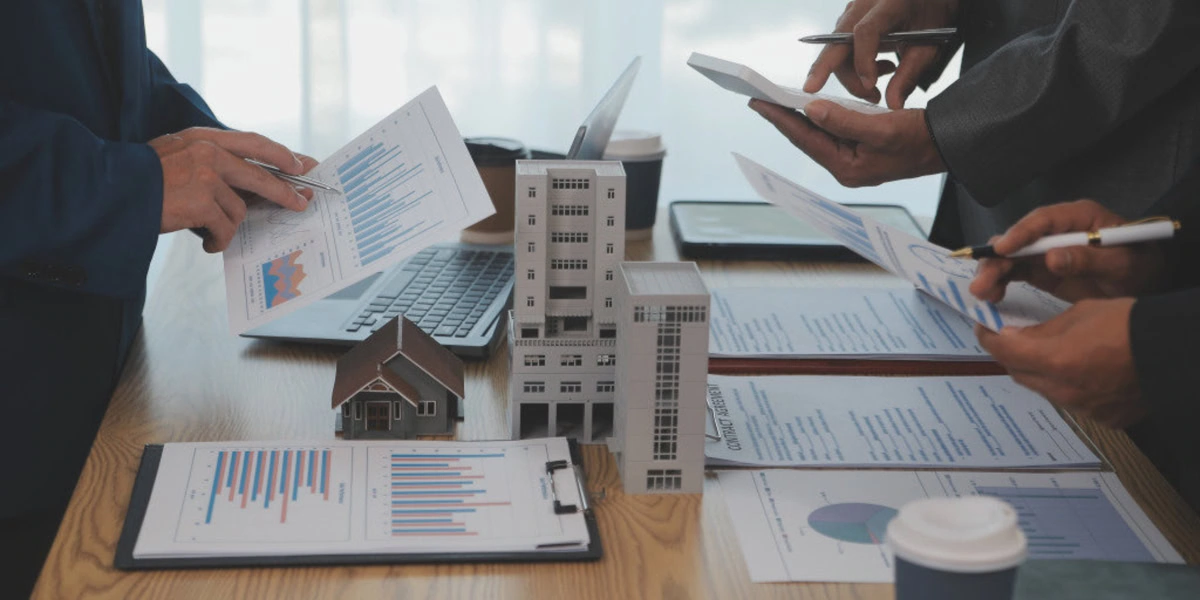 People examining documents and charts around a table with model buildings and coffee cups.