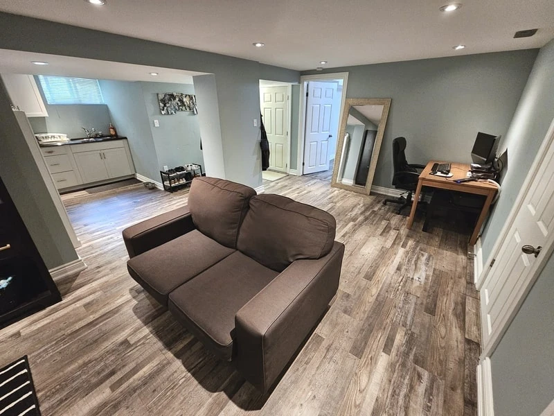 A basement room featuring a small seating area with a brown loveseat, a wooden desk with a computer, a full-length mirror, and a kitchenette in the background. The flooring is wooden—a perfect space managed by top-notch property management services near me.