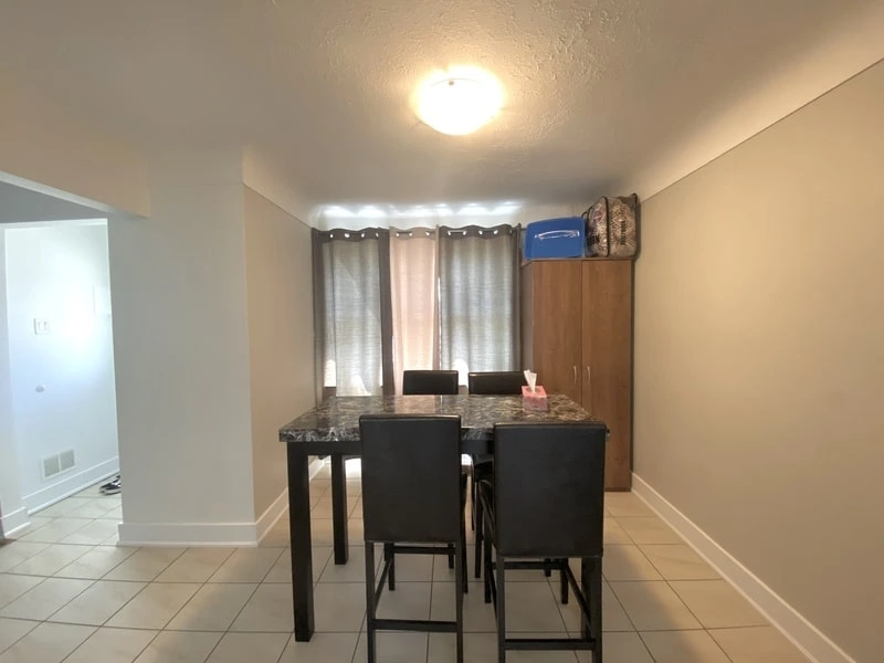 A dining room with a dark wooden table, four black chairs, a tissue box, and a wooden cabinet with luggage on top. Grey curtains cover the window and the flooring is tiled. Ceiling light is on. Ideal for those seeking property management services near me, offering comfort and style in every corner.