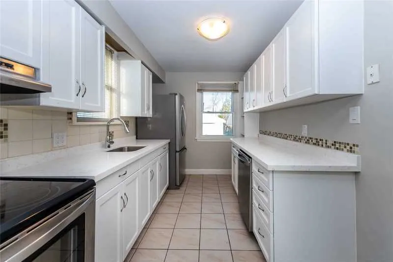 A kitchen with white cabinetry, stainless steel appliances, tiled floor, and a window providing natural light at the end of the room is an inviting space. For top-notch property management services near me, this beautifully designed kitchen is a perfect example of refined taste and functionality.