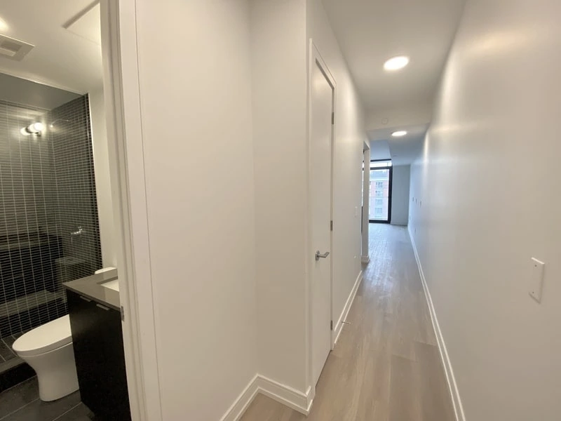 A long, narrow hallway with a wooden floor leading to a brightly lit room. On the left, a sleek, modern bathroom with dark tile walls and a white toilet is visible. For those seeking top-notch property management services near me, this space reflects impeccable standards.