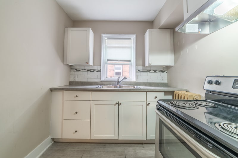 A well-maintained kitchen features white cabinets, a stainless steel sink under a window, and a stainless steel stove with black coils on the right side. Wall-mounted white cabinets are above the sink area, showcasing the care taken by a good property management company.