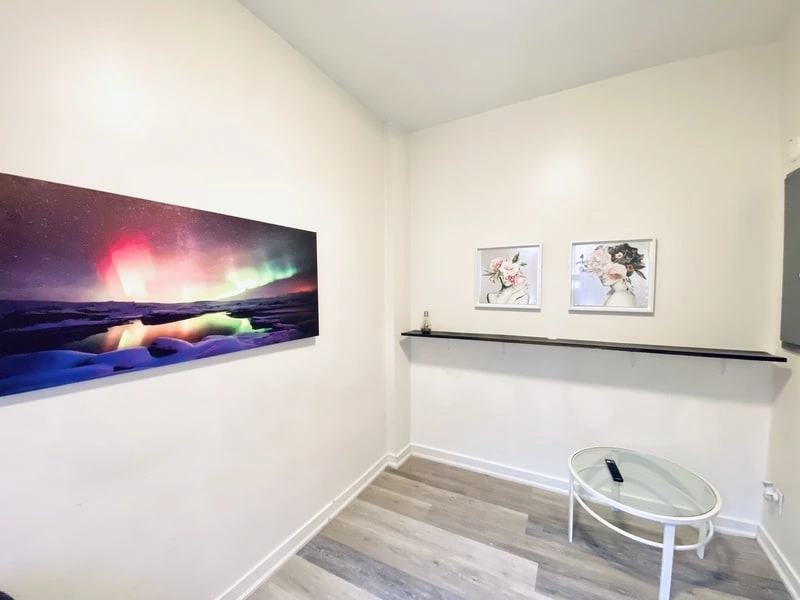 A small room with white walls features a large photo of an aurora, two small framed pictures of flowers on a shelf, and a round glass table holding a remote control. The laminate floor has light wood patterning, showcasing the attention to detail you'd expect from a good property management company.