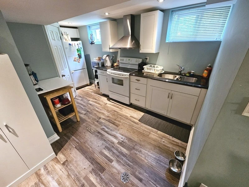 A compact kitchen with light gray walls and wooden flooring, featuring a fridge, stove, microwave, sink, white cabinets, and a countertop. A dog bowl is on the floor near the sink area. This well-maintained space reflects the care of a good property management company.