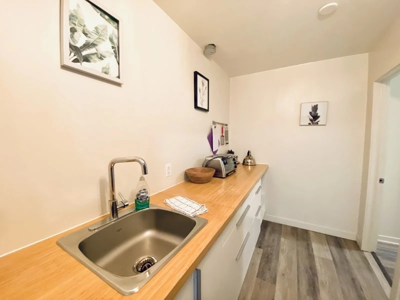 A small kitchen with a wooden countertop, sink, and various appliances. Three framed pictures adorn the wall, while the floor boasts a wood-like finish—a testament to the care and detail that a good property management company ensures.