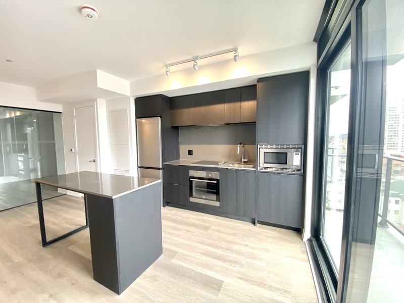 A modern kitchen with dark cabinets, stainless steel appliances, an island with a gray countertop, and large windows with city views – all meticulously maintained by a good property management company ensuring top-tier upkeep and service.