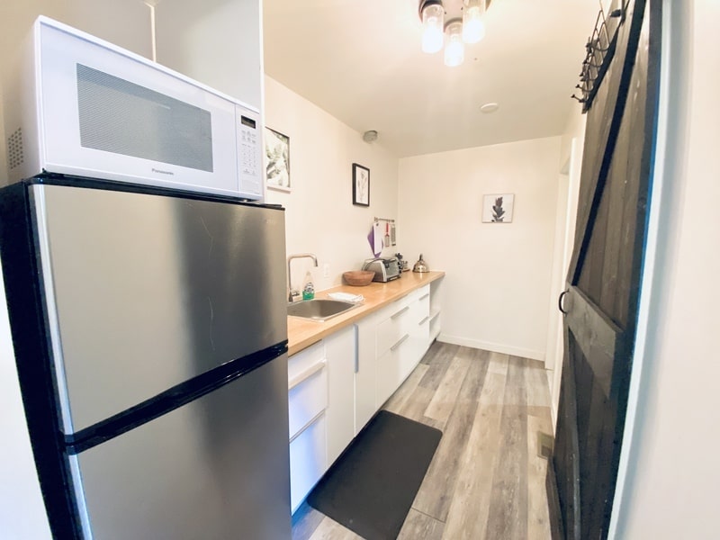 A compact kitchen with a refrigerator, microwave, sink, and wooden countertops. The space has white cabinets, a dark mat, and framed pictures on the walls—indicating the touch of a good property management company ensuring everything is in perfect order.