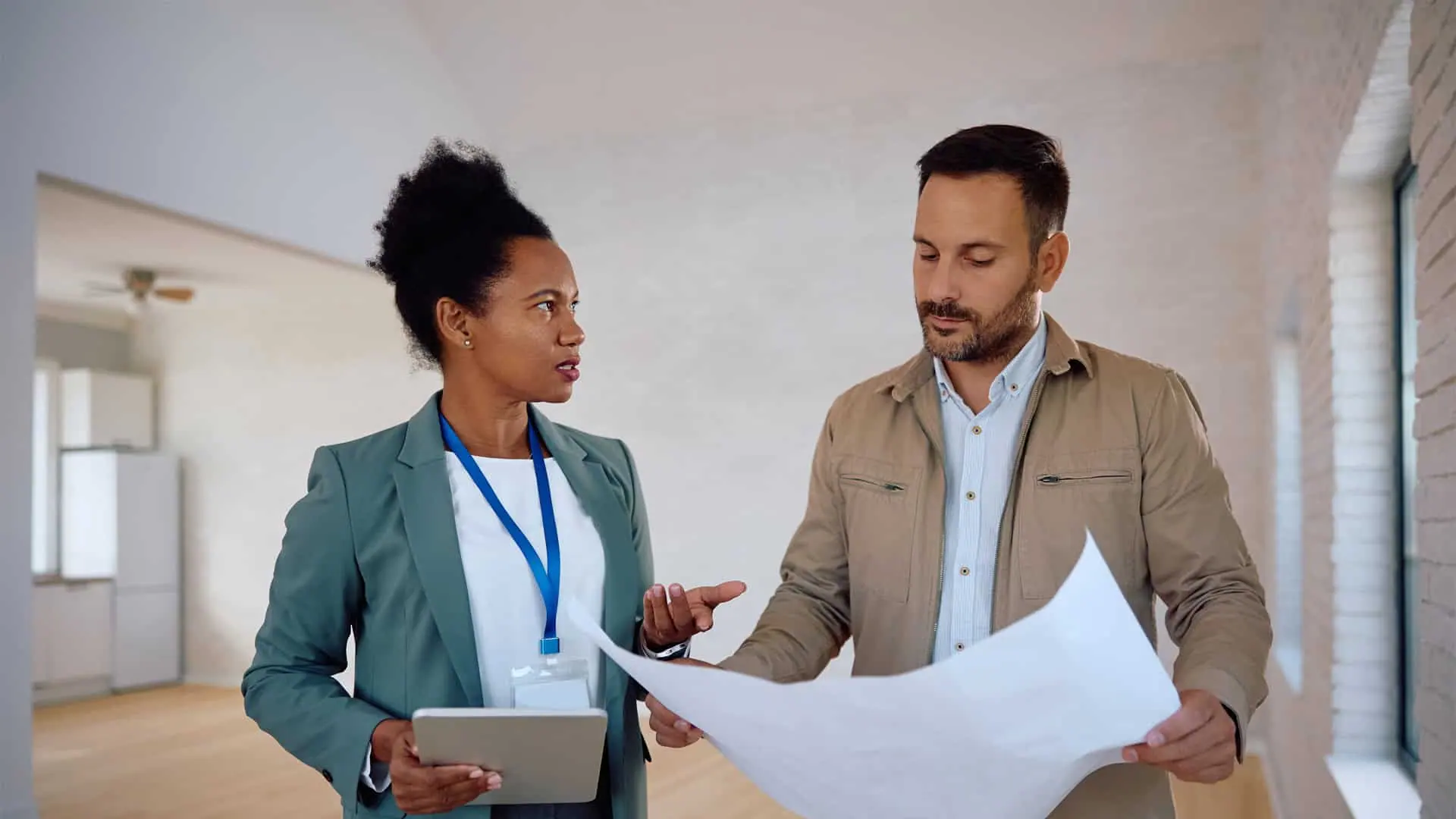 Two people stand in an empty room managed by Hamilton Property Managers, discussing a large paper document. One holds a tablet while the other reviews the document.