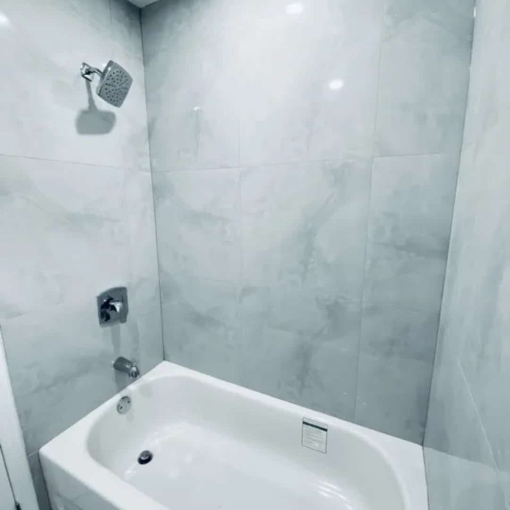 A white bathtub with chrome fixtures is set against a wall of large gray marble tiles, featuring a square showerhead—a luxurious touch often highlighted by property management agencies.