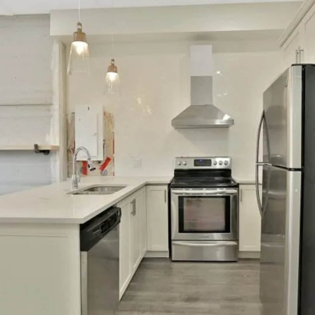 This modern kitchen, managed by a leading property management company, features stainless steel appliances, white cabinetry, and wooden flooring. Enjoy amenities like an oven, dishwasher, refrigerator, sink, and stylish pendant lights in this meticulously maintained space.