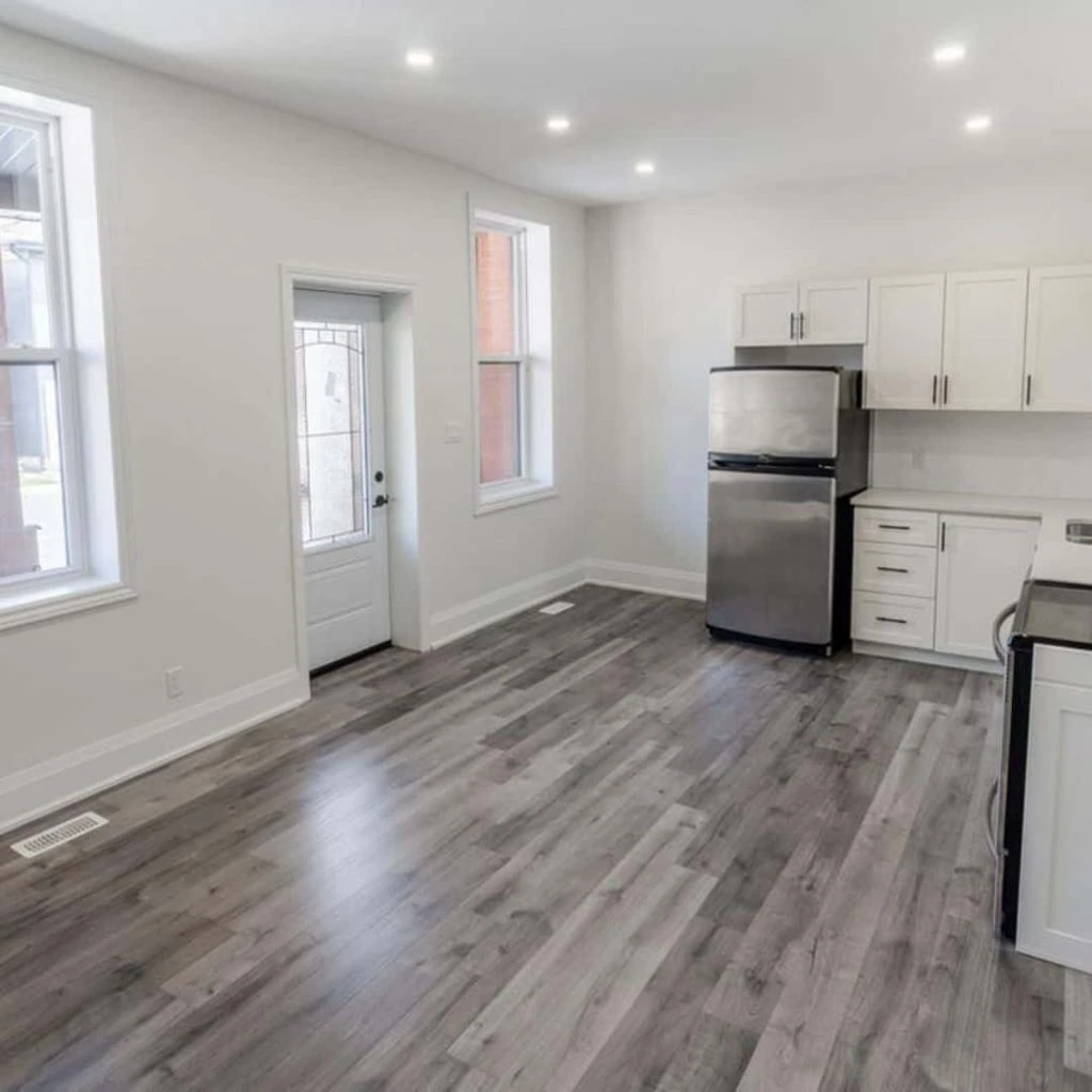 Modern kitchen with light gray wood flooring, white cabinets, and stainless steel appliances, expertly maintained by our Rent Management Company. Two large windows and a glass-paneled door flood the space with natural light.