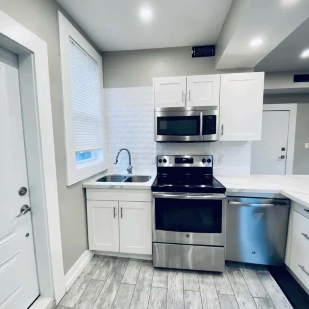 Modern kitchen with white cabinets, stainless steel appliances, a sink under a window, and light gray flooring—perfectly maintained by top rental property management companies.