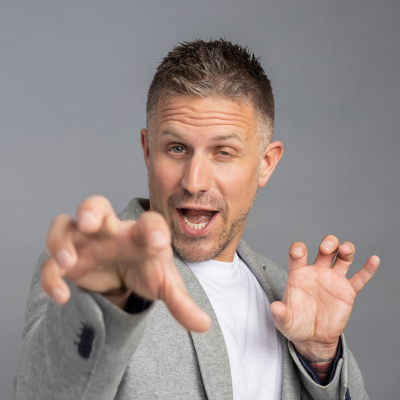 A man in a gray blazer and white shirt poses playfully with a hand extended forward, making a claw-like gesture against a plain background, embodying the dynamic spirit of Hamilton Companies—known for property management excellence.