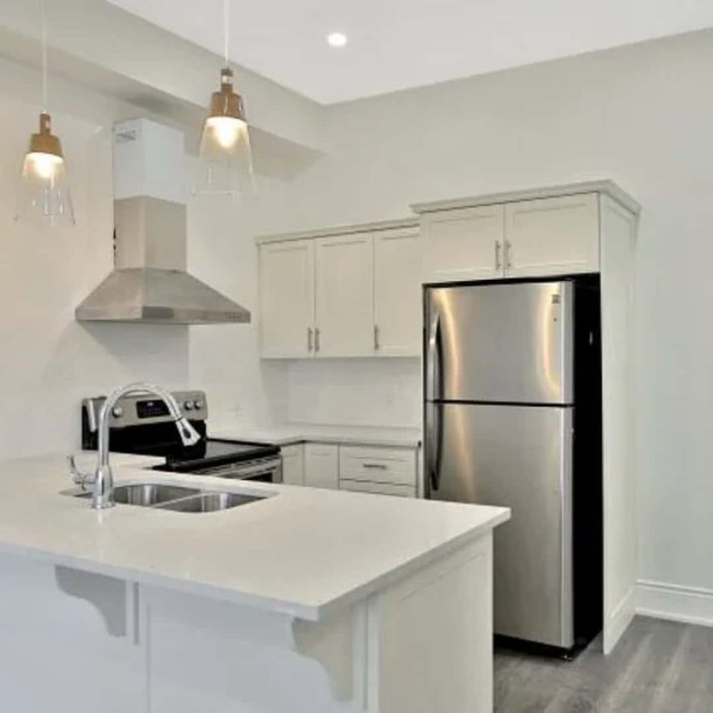 Modern kitchen managed by a top-notch property management company, featuring white cabinets, a stainless steel refrigerator, oven, and range hood. The center island includes a sink with stylish hanging pendant lights overhead.