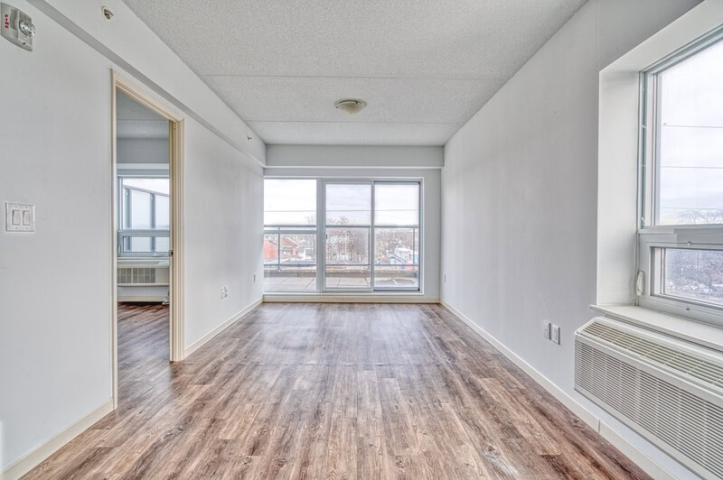 Empty room with large windows, light gray walls, and wooden floors. A small air conditioning unit is installed below the window. Managed by a top-tier property management company, it features a doorway that leads to an adjacent room, property managers in Hamilton Ontario