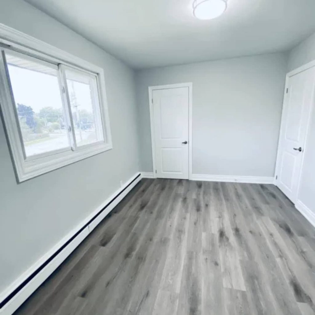 The empty room, managed by leading property management agencies, features light gray walls, wood-look flooring, a window filling the space with natural light, and two closed doors.