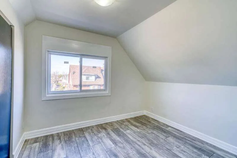 A small, empty room with light-colored walls, a sloped ceiling, wooden flooring, and a window offering a view of neighboring houses. Perfectly maintained by the property management company, property managers in Hamilton Ontario