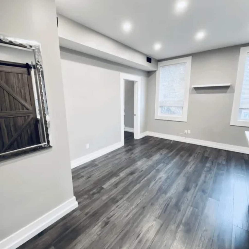 A clean, vacant room with light gray walls and dark wood flooring, perfect for showcasing by rental property management companies. Featuring a large window, a wall-mounted shelf, and a decorative mirror beside the door, it's an inviting space ready for new possibilities.