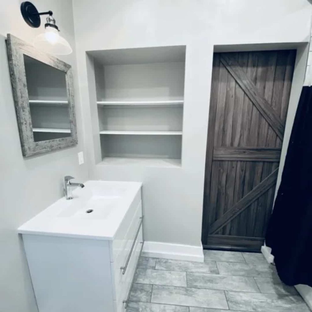 This cozy bathroom, ideal for rental property management companies, features a white vanity, mirror, open shelves, and a charming wooden barn door. The gray floor tiles and wall-mounted light fixture add a touch of elegance.