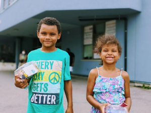 Two children are smiling brightly outside a building, each holding a small package. The boy, wearing a green shirt with text, and the girl in her colorful dress seem to embody the spirit of giving back in their community's found spaces.