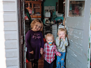 Three children stand in a doorway, part of a Found Spaces program aimed at giving back to the community. The girl on the left wears a purple dress, while the two toddlers to her right wear casual outfits—one holding a pacifier. They smile at the camera, radiating joy and innocence.