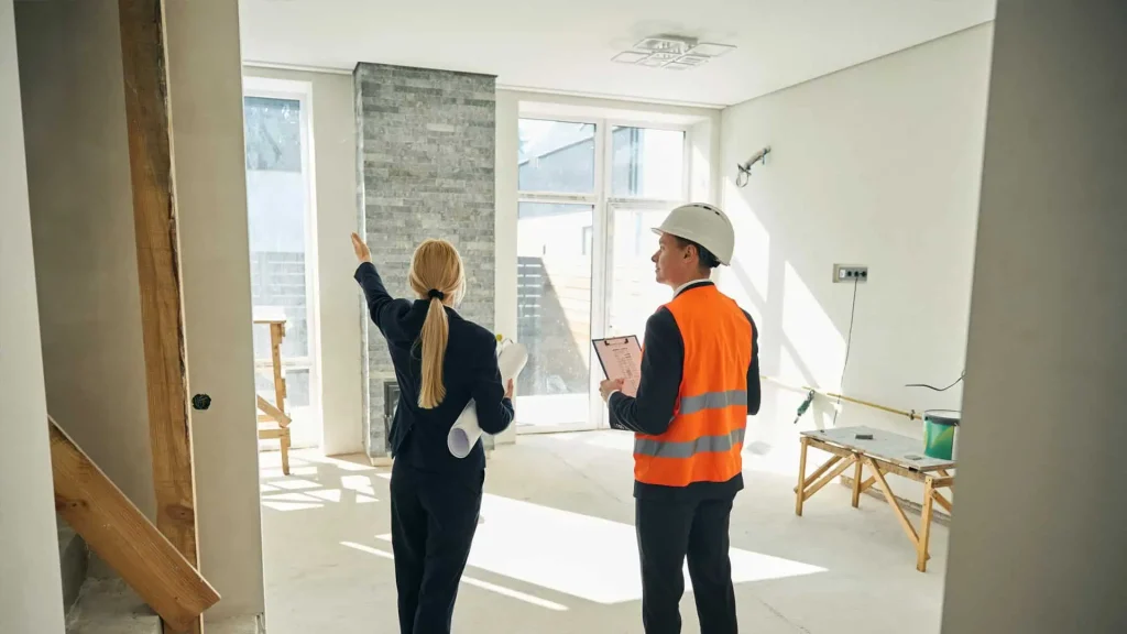 Two people in a partially renovated room, one in a suit discussing property management strategies and the other in a hard hat and safety vest holding a clipboard, keenly noting details for the landlords.