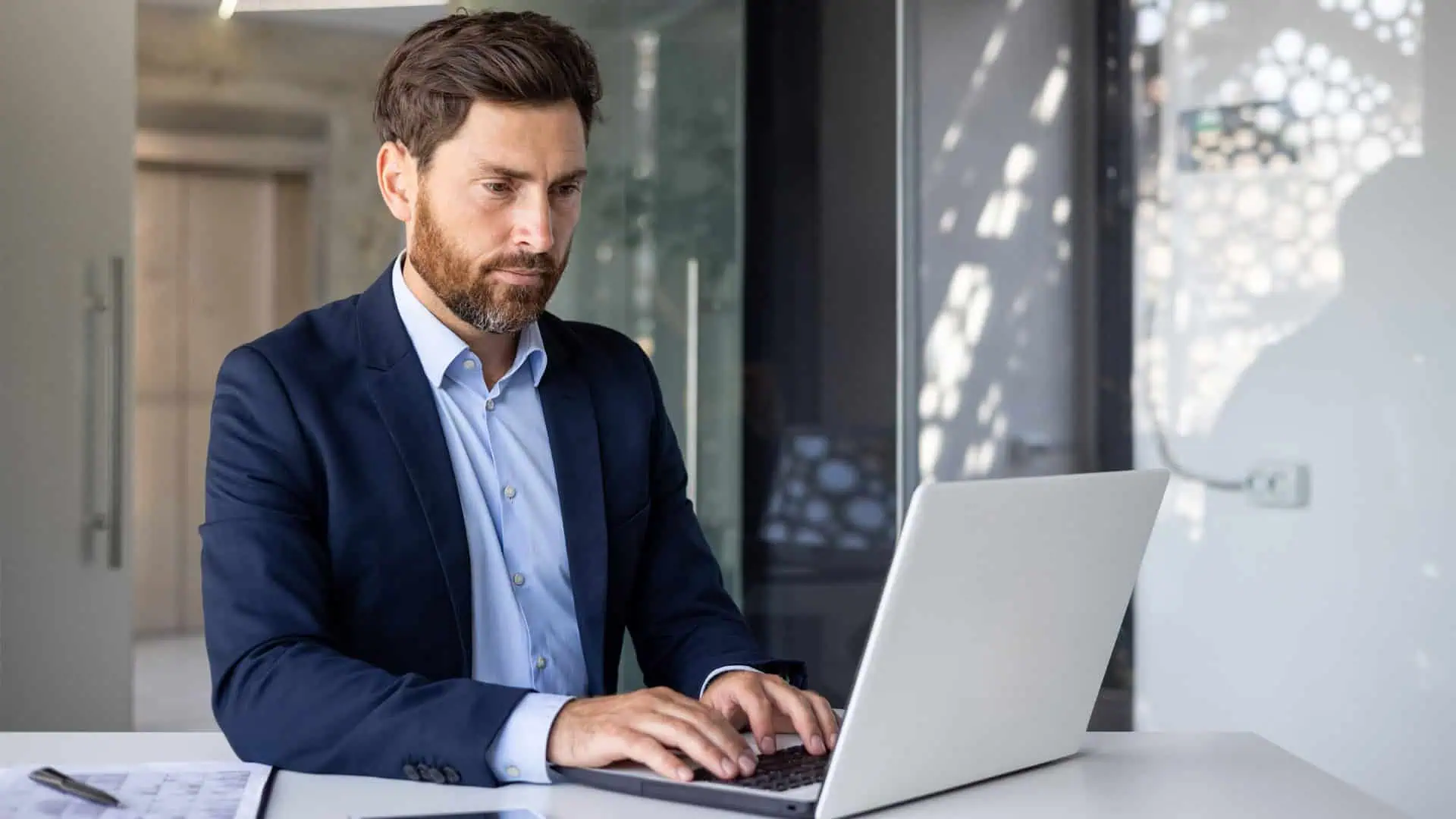 A man in a suit focuses intently as he types on a laptop at his desk, orchestrating multi-property management services seamlessly in the bustling office setting, multi property management services