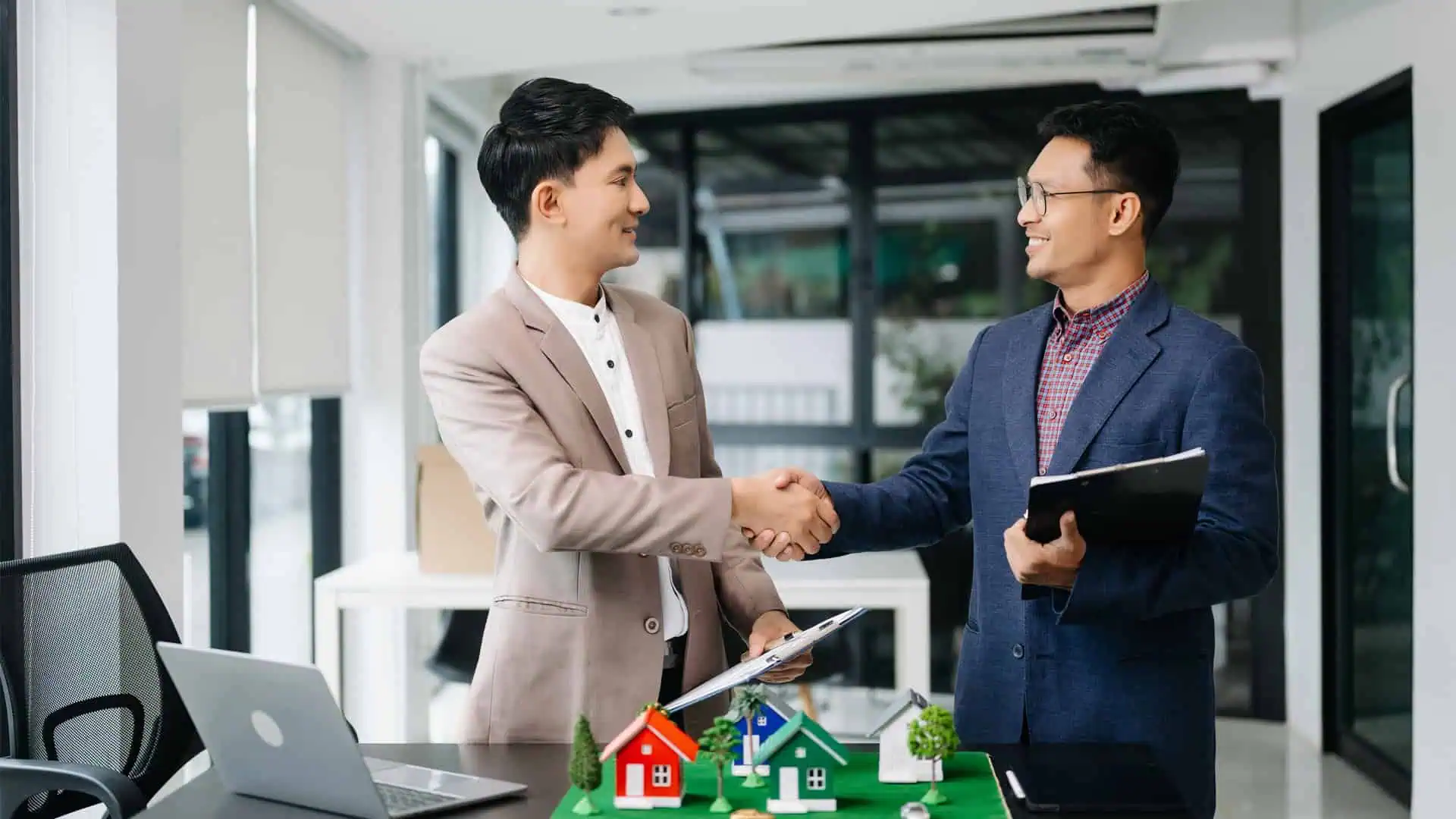 Two men in suits shake hands in a modern Hamilton office, sealing a property investment deal. A model of houses sits on the table between them, Property Investment Hamilton